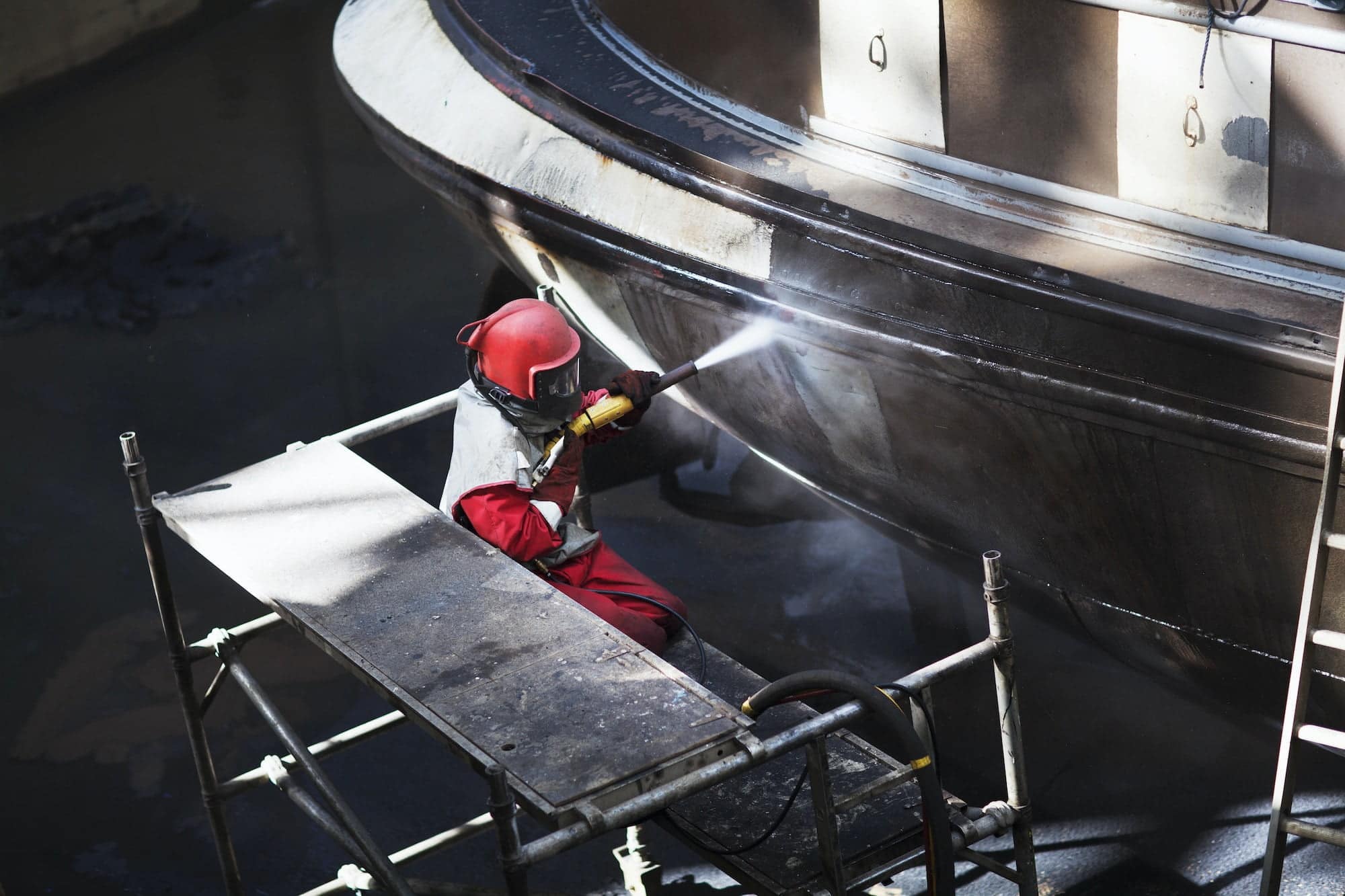 Worker cleaning hull of boat with high pressure hose in shipyard