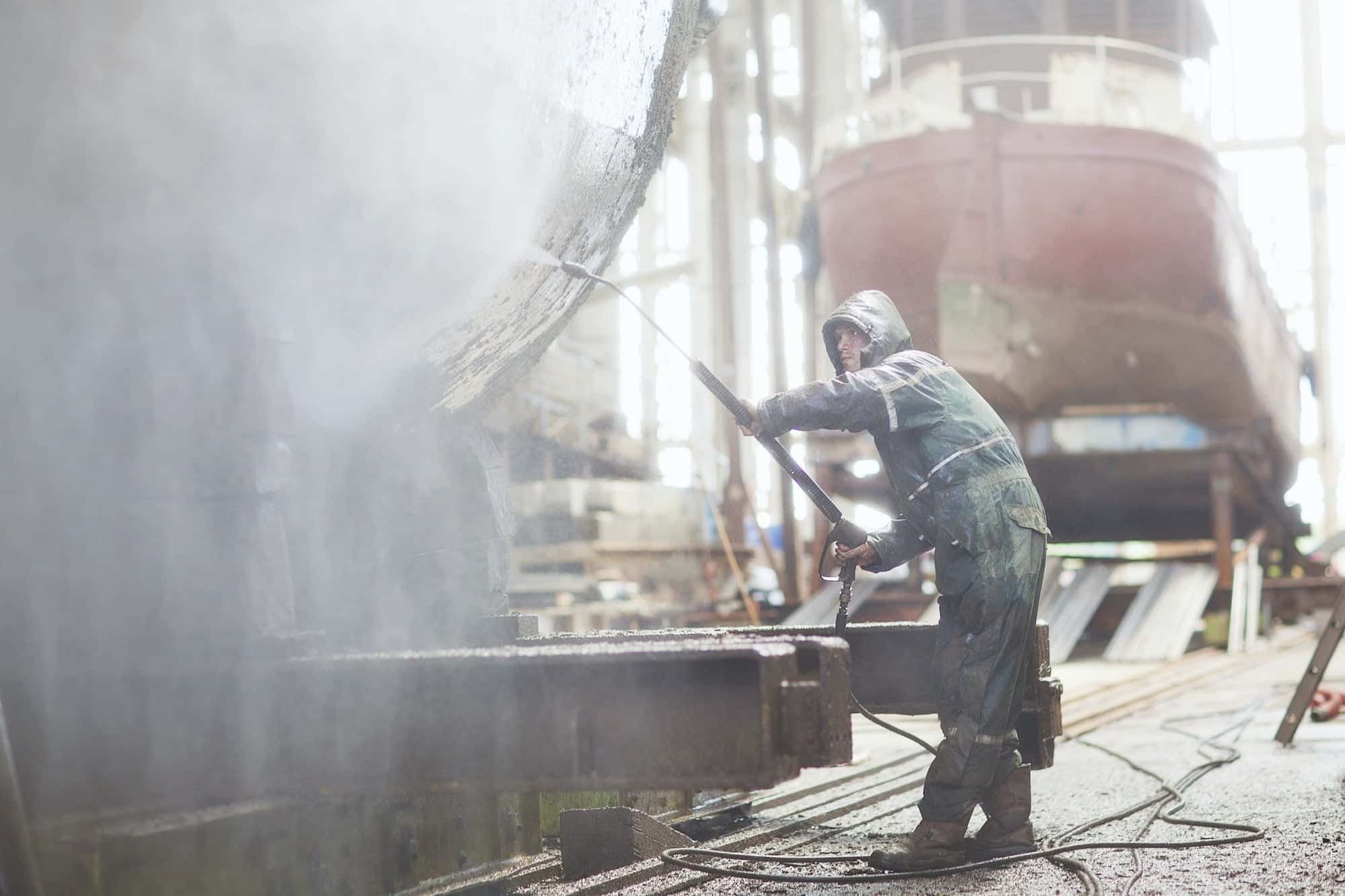 Worker using high pressure hose on boat in shipyard workshop